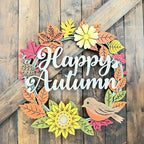 A decorative autumn wreath sign with layered paper cutout leaves, flowers, a bird, and the words “Happy Autumn” in white script, displayed against a rustic wooden plank background.