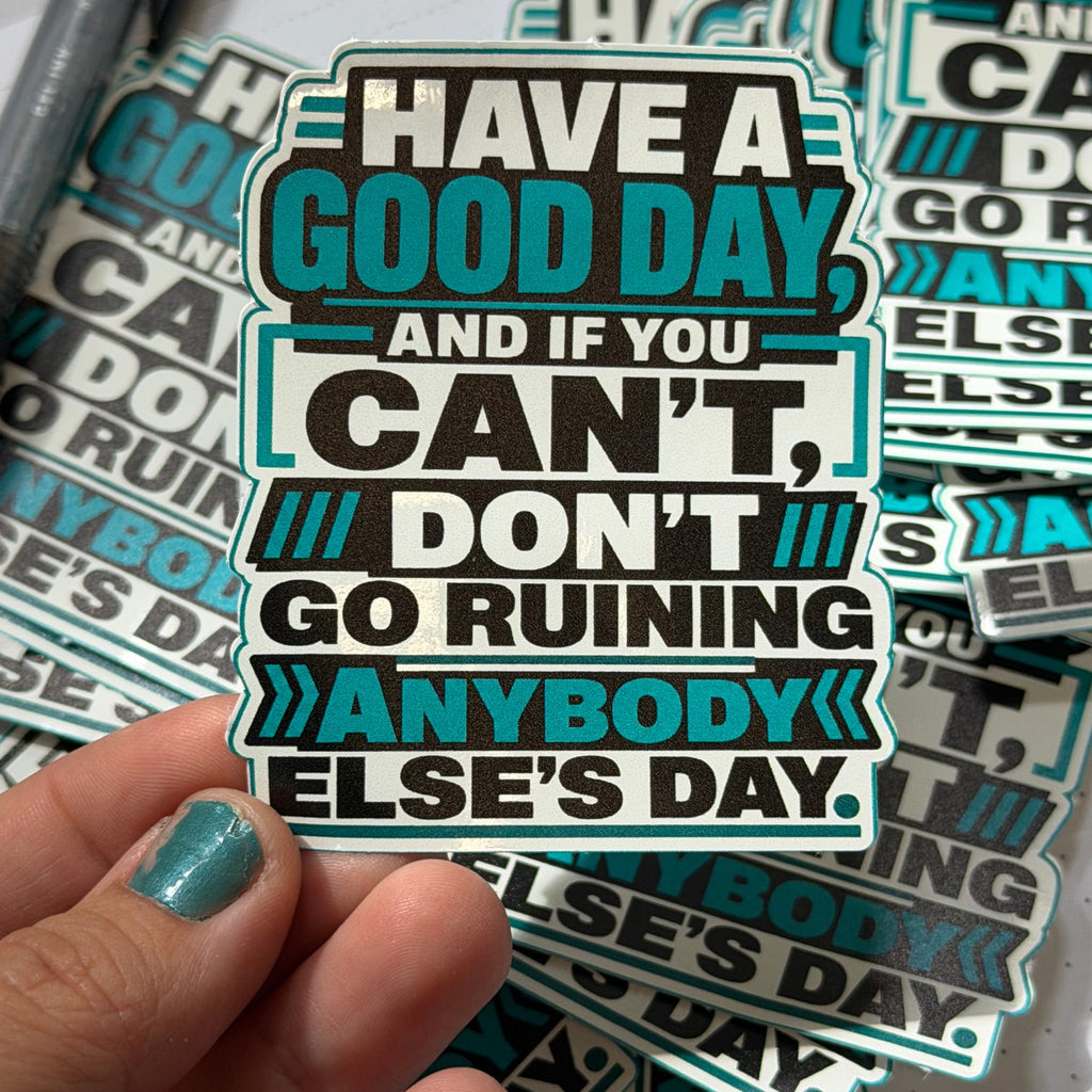 A hand with teal nail polish holds a die-cut sticker that reads 'HAVE A GOOD DAY, AND IF YOU CAN'T, DON'T GO RUINING ANYBODY ELSE'S DAY.' Multiple identical stickers are stacked in the background on a white surface.