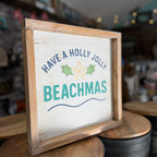 A rustic wooden framed sign with a whitewashed face that reads 'HAVE A HOLLY JOLLY BEACHMAS' in navy and teal lettering with green holly leaves and a yellow star, displayed on wooden stools in a shop setting.