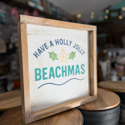 A rustic wooden framed sign with a whitewashed face that reads 'HAVE A HOLLY JOLLY BEACHMAS' in navy and teal lettering with green holly leaves and a yellow star, displayed on wooden stools in a shop setting.