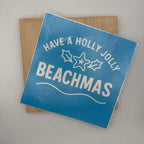 A square blue sign with white text reading 'HAVE A HOLLY JOLLY BEACHMAS' and simple holiday graphics, photographed resting on a light wood board against a plain light background.