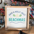 A square wooden framed sign with a distressed white background that reads 'HAVE A HOLLY JOLLY BEACHMAS' in blue and teal lettering, with a starfish and green leaves graphic, displayed on a wooden table in a shop setting.