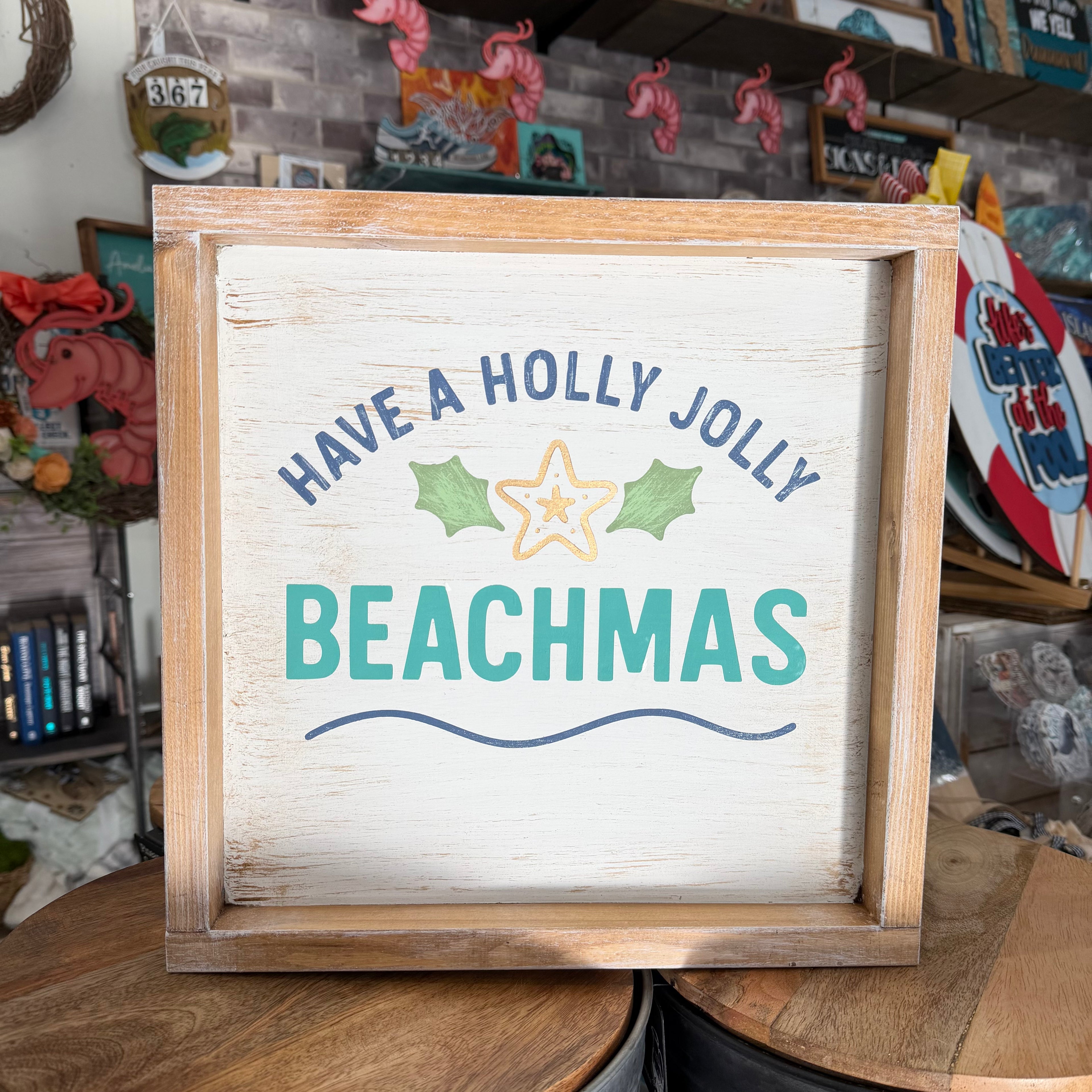 A square wooden framed sign with a distressed white background that reads 'HAVE A HOLLY JOLLY BEACHMAS' in blue and teal lettering, with a starfish and green leaves graphic, displayed on a wooden table in a shop setting.