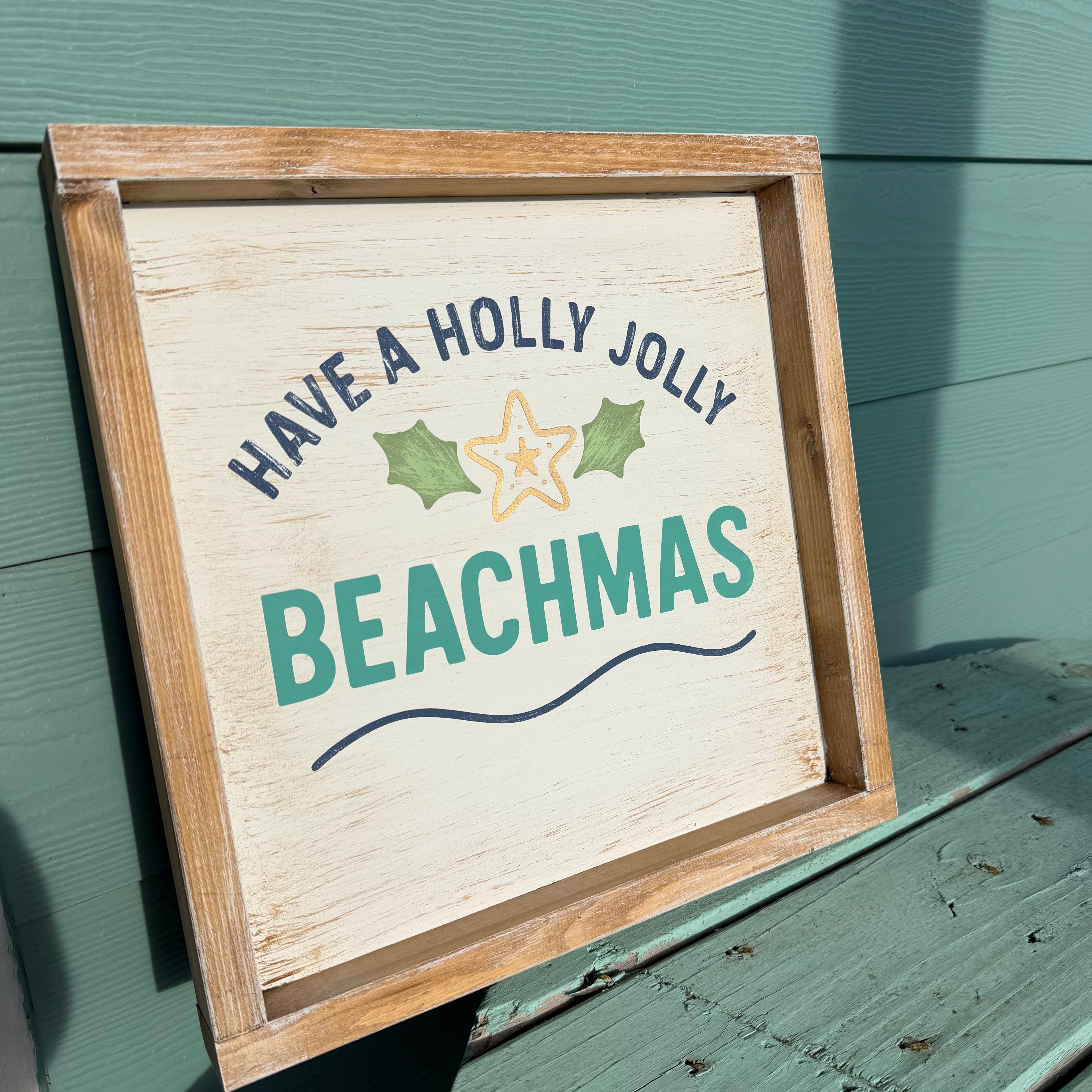 A rustic wooden framed sign with a distressed cream face, reading “HAVE A HOLLY JOLLY BEACHMAS” in navy and teal lettering with a starfish and holly graphic, leaning against a painted green wooden wall.