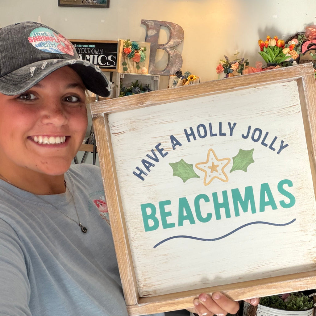 A smiling woman in a gray t-shirt and baseball cap holds up a rustic white wooden sign that reads 'HAVE A HOLLY JOLLY BEACHMAS' with green holly leaves, a yellow star, and blue wavy lines. The background shows a decorated indoor space with a large letter 'B' and artificial flowers.