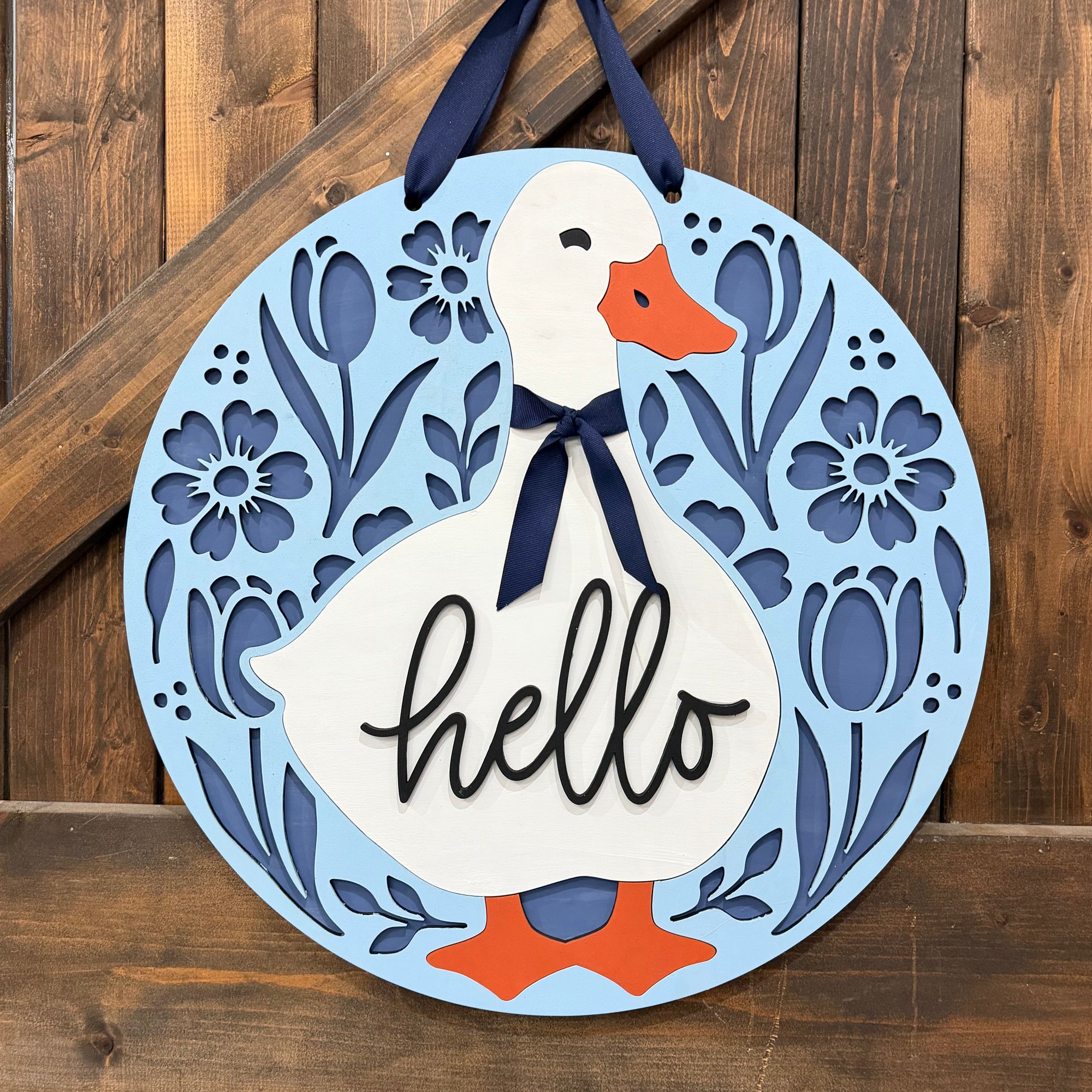 Round decorative wall hanging with a white duck illustration, blue floral cutout border, orange beak and feet, navy hanging ribbon, and the word "hello" in black script on a light blue background, shown against a wooden plank wall.