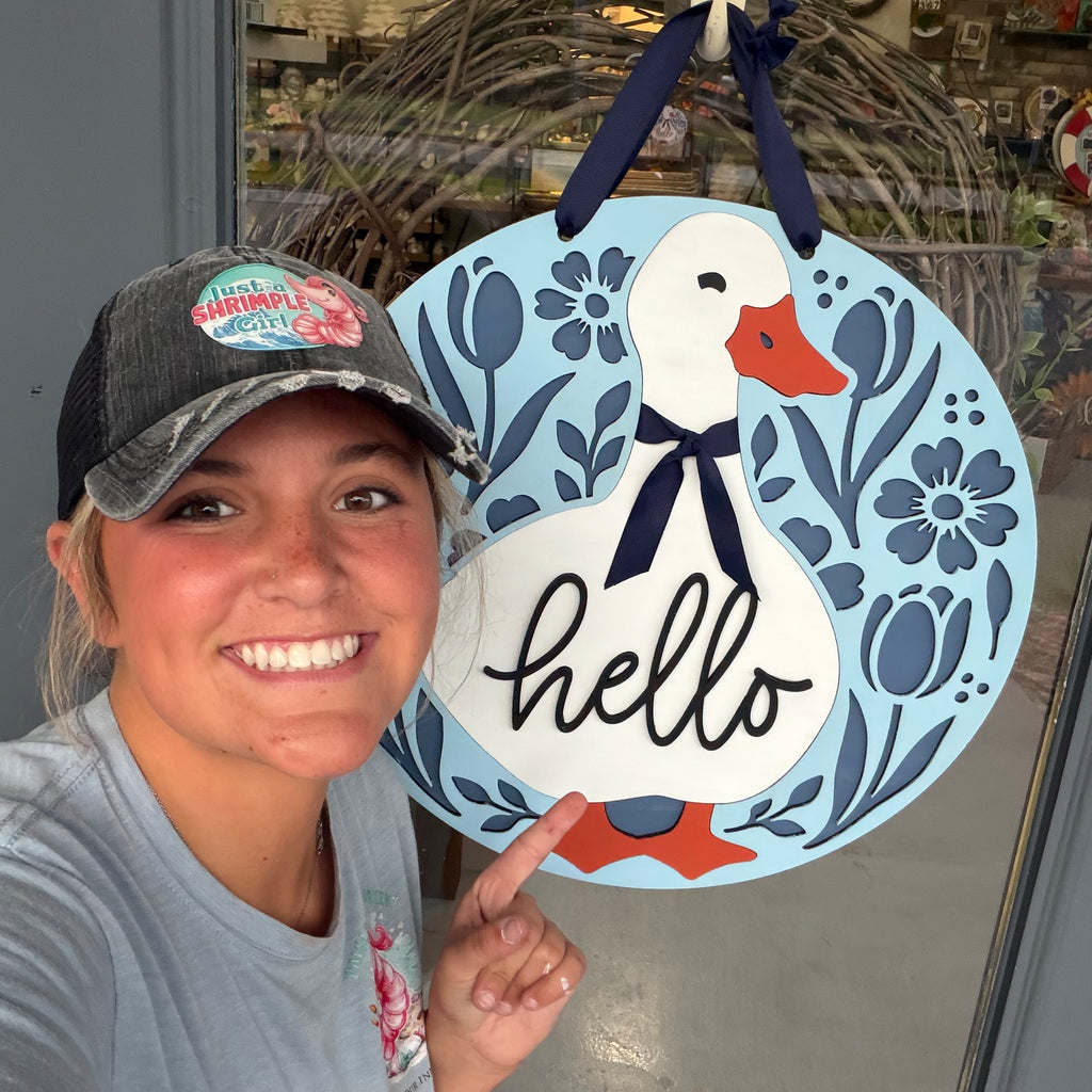A smiling woman wearing a gray cap points to a decorative hanging sign shaped like a white duck with an orange beak, blue floral border, and the word 'hello' in black script. The sign is displayed in a storefront setting.