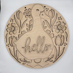 A round, laser-cut wooden wall sign featuring a duck silhouette with the word 'hello' cut into the center, surrounded by tulips and flowers. The natural wood grain is clearly visible against a plain white background.