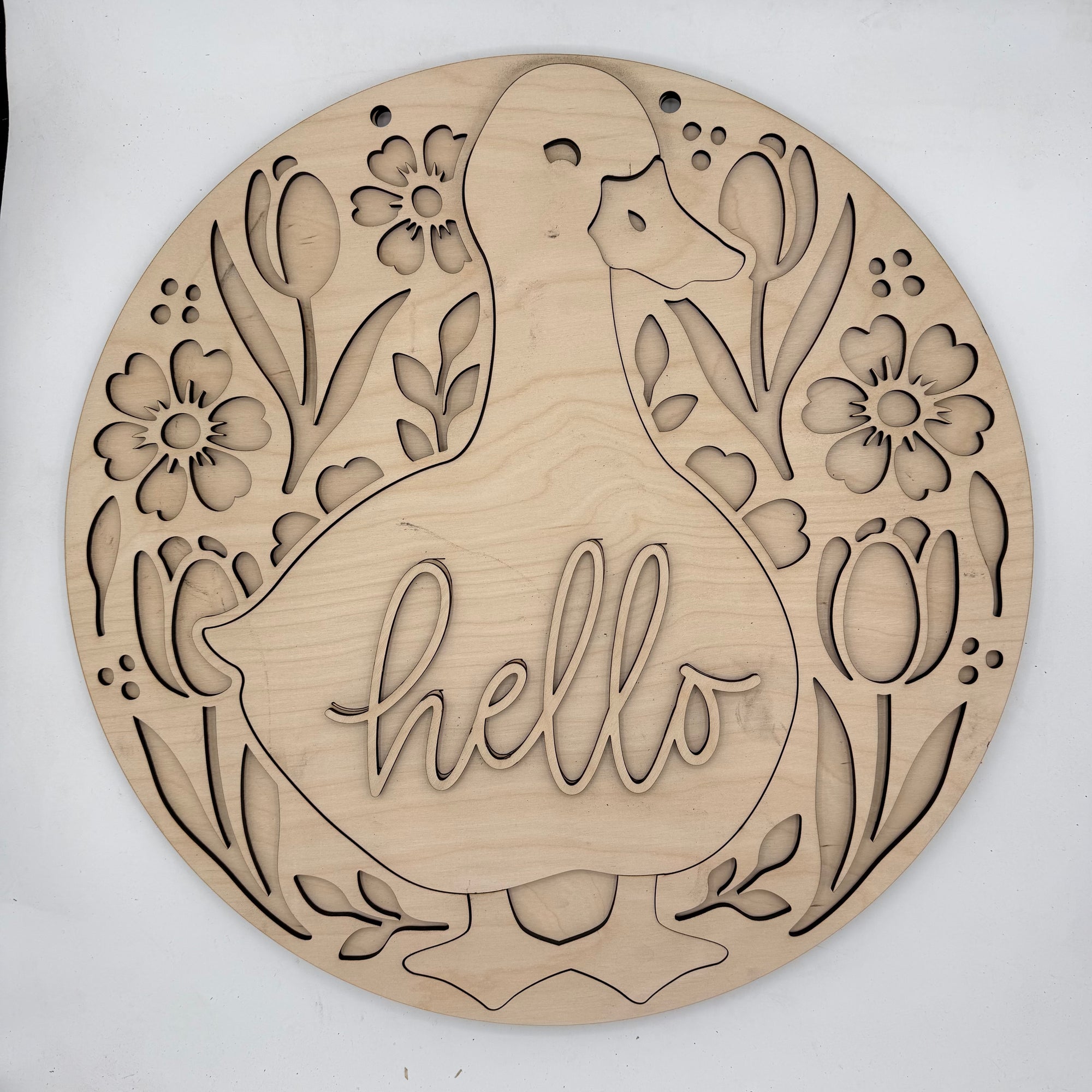 A round, laser-cut wooden wall sign featuring a duck silhouette with the word 'hello' cut into the center, surrounded by tulips and flowers. The natural wood grain is clearly visible against a plain white background.