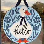 A round decorative hanging sign featuring a white duck with an orange beak and feet, a navy ribbon bow, and the word "hello" in black script. The sign is light blue with navy floral and leaf cutout designs around the border, hanging on a glass door or window.