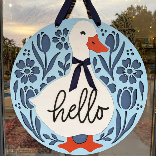 A round decorative hanging sign featuring a white duck with an orange beak and feet, a navy ribbon bow, and the word "hello" in black script. The sign is light blue with navy floral and leaf cutout designs around the border, hanging on a glass door or window.