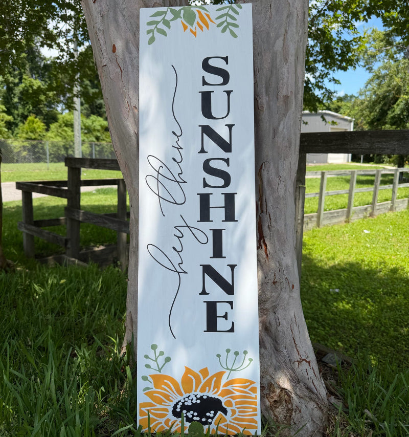 Decorative sign with 'SUNSHINE' and sunflower design attached to a tree outdoors.