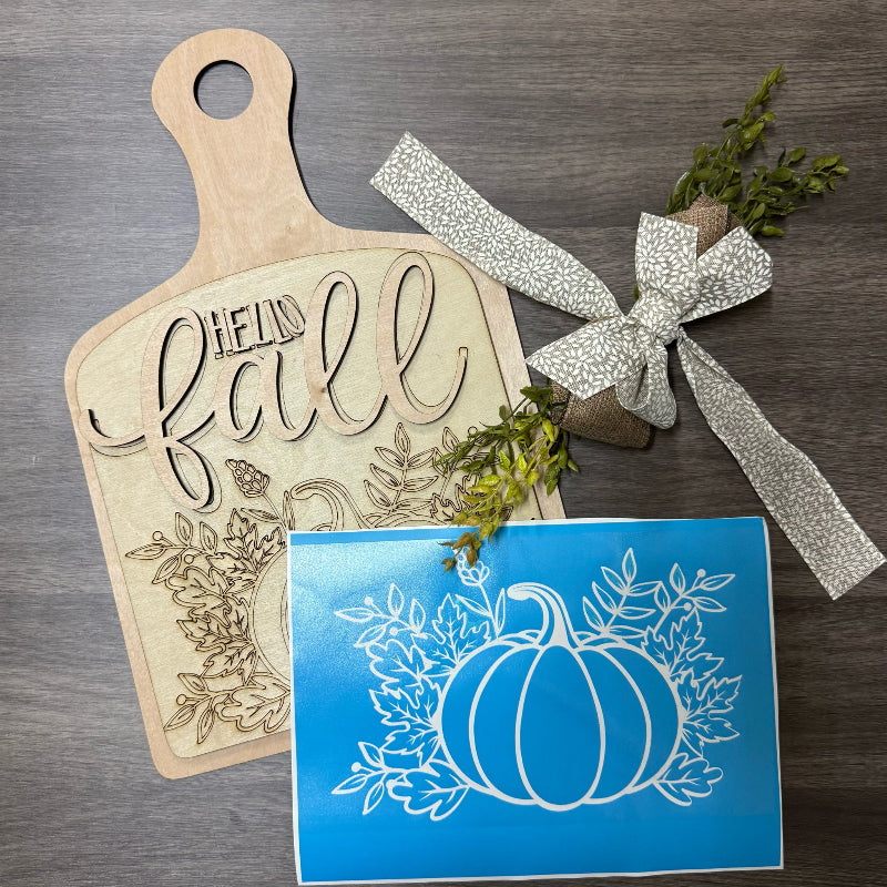 Wooden cutting board with 'Hello Fall' engraving, decorative ribbon, and pumpkin design card on a wooden surface.