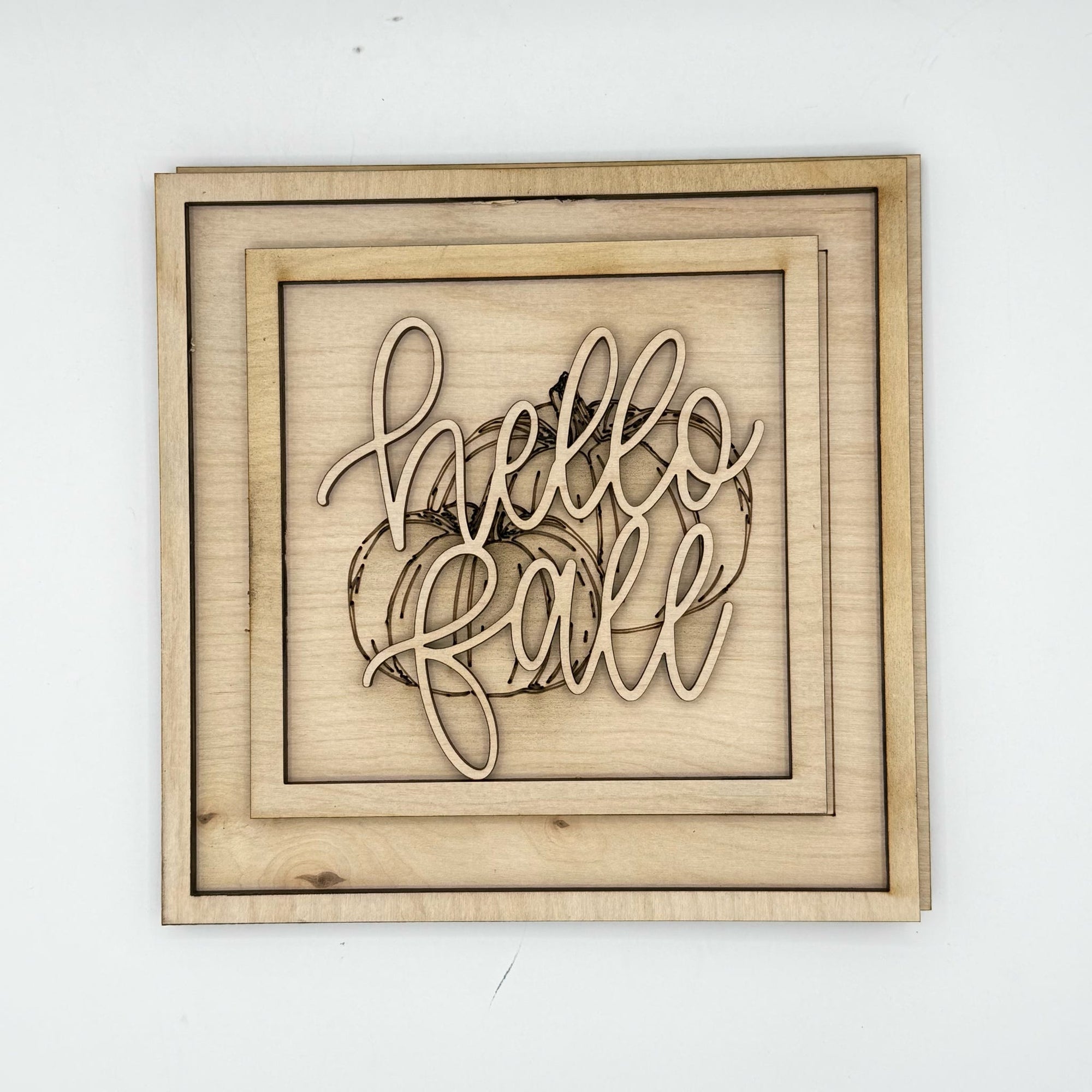 A square wooden wall sign with layered natural wood frames and laser-cut script reading 'hello fall' over a pumpkin outline, shown against a plain white background.