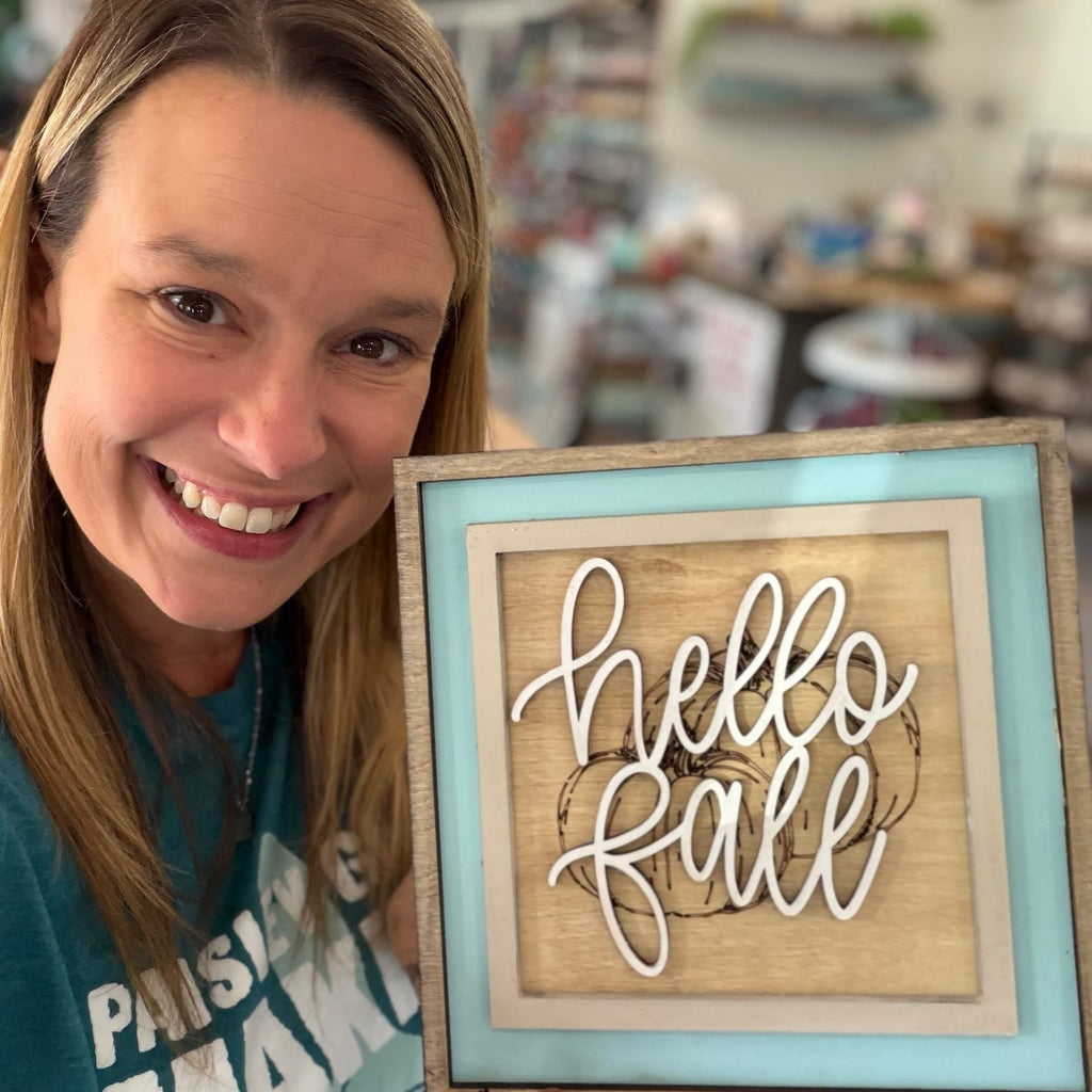 A smiling woman holds up a framed decorative sign that reads 'Happy Fall' in white script over a light wood background with layered turquoise and beige frames. The background shows a softly blurred craft shop interior with shelves and string lights.