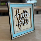 A square decorative wooden sign with layered frames in gray, light blue, and natural wood, featuring a laser-cut white script 'hello fall' design on a light wood background, set on a brown surface with a blurred indoor background.