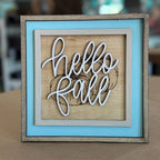 A square framed decorative sign with layered wood tones, a light blue inner frame, and white script lettering that reads 'hello fall' over a rustic wood background.