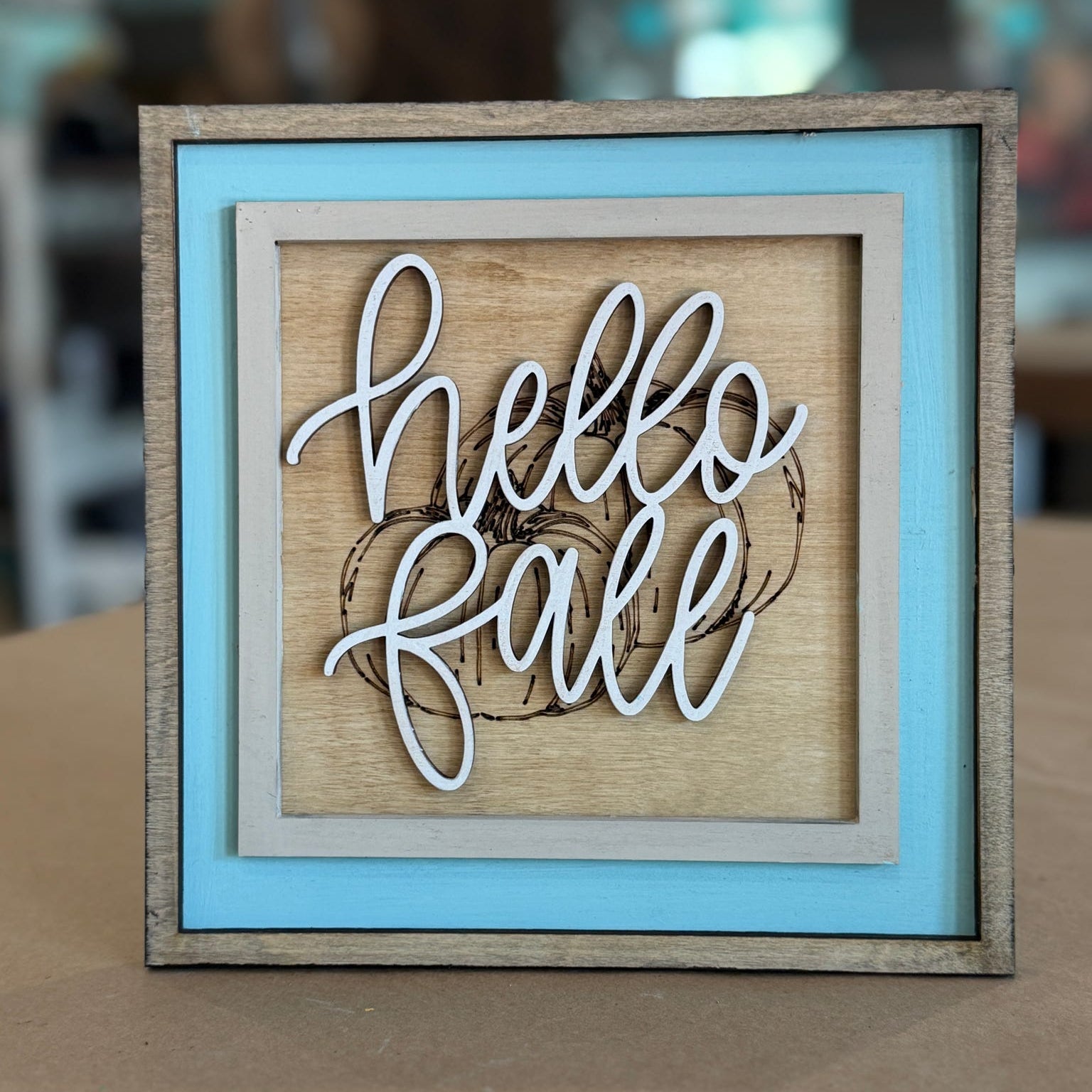 A square framed decorative sign with layered wood tones, a light blue inner frame, and white script lettering that reads 'hello fall' over a rustic wood background.