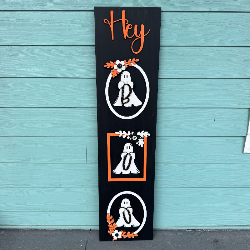 Decorative Halloween sign with ghosts and floral elements on a black background against a blue wooden wall.