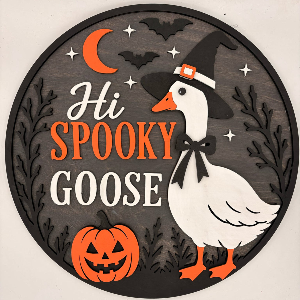 A round Halloween wall sign featuring a white goose wearing a witch hat, orange text reading 'Hi SPOOKY GOOSE,' a jack-o'-lantern, bats, stars, and a crescent moon on a dark gray background.