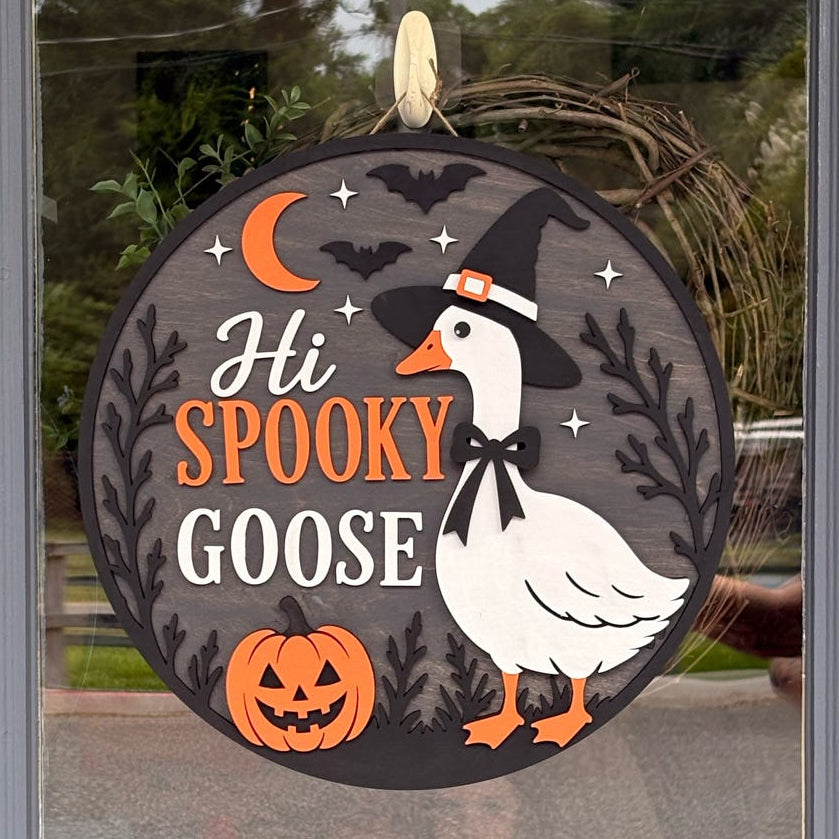 A round Halloween door hanger with the text 'Hi SPOOKY GOOSE' and an illustration of a white goose wearing a witch hat, orange pumpkins, bats, and a crescent moon, displayed on a glass door.
