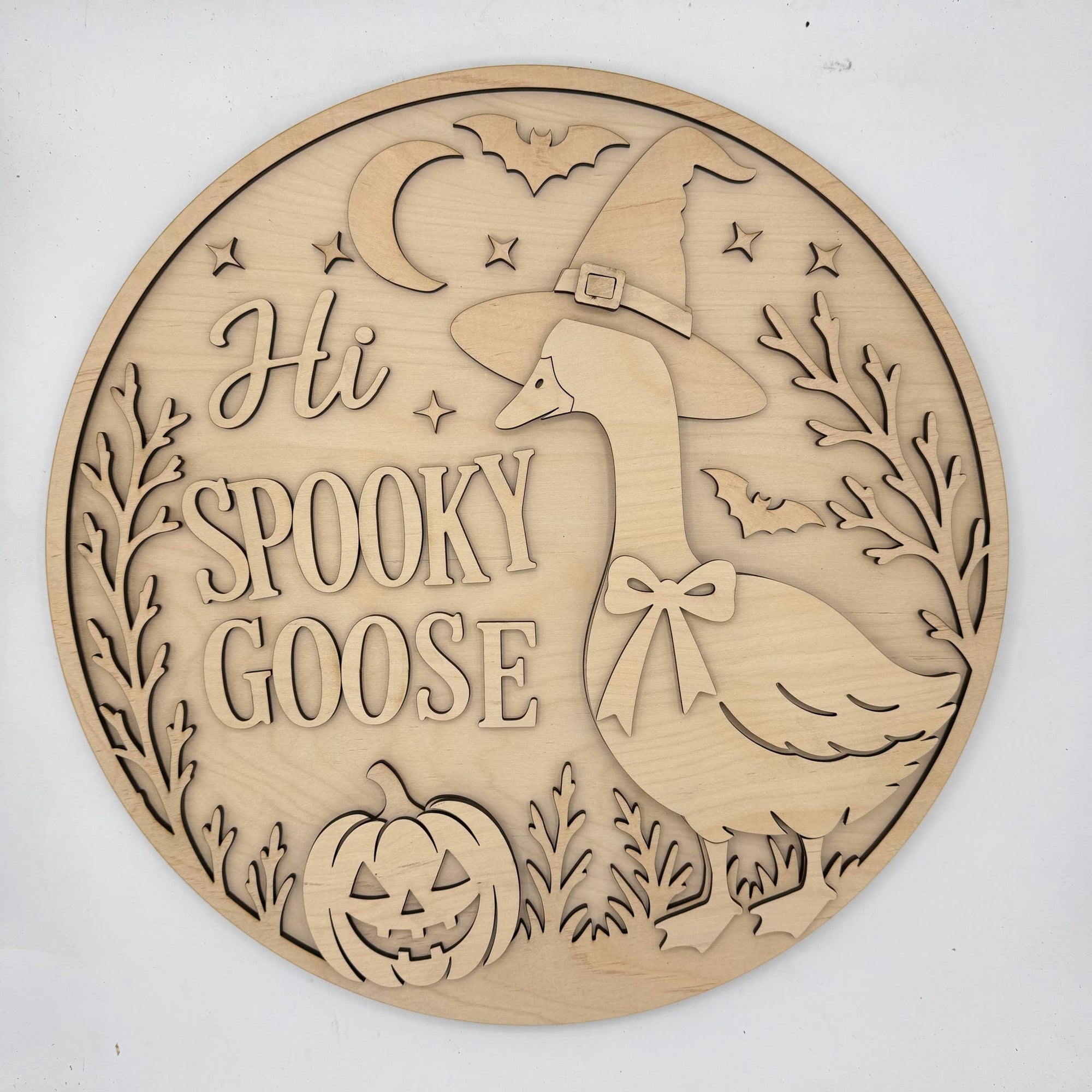 Round laser-cut wooden plaque featuring a carved goose wearing a witch hat, the phrase 'Hi SPOOKY GOOSE,' a jack-o'-lantern, bats, stars, and a crescent moon on a light wood background.