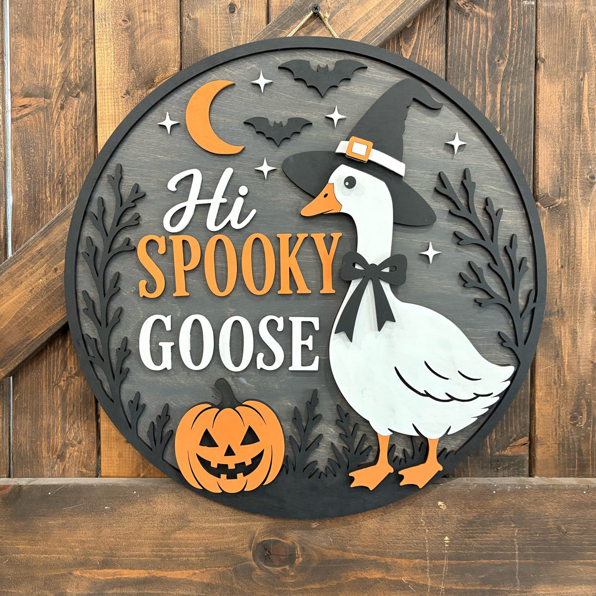 Round wooden Halloween wall sign featuring a white goose wearing a witch hat, orange text reading 'Hi SPOOKY GOOSE,' a jack-o'-lantern, bats, stars, and a crescent moon on a rustic wood plank background.