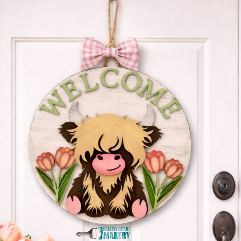 Round wooden sign with a cartoon Highland cow, tulips, and the word 'Welcome' on a white door.