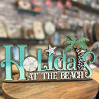 A decorative wooden sign reading 'Holidays at the Beach' in turquoise and white lettering, with a green palm tree and sand dollar accents, displayed on a wooden surface with a blurred shop background.