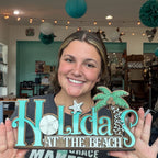 A smiling woman holds a decorative wooden sign that reads 'Holidays at the Beach' with turquoise lettering, a palm tree, and starfish motifs in a craft store setting.