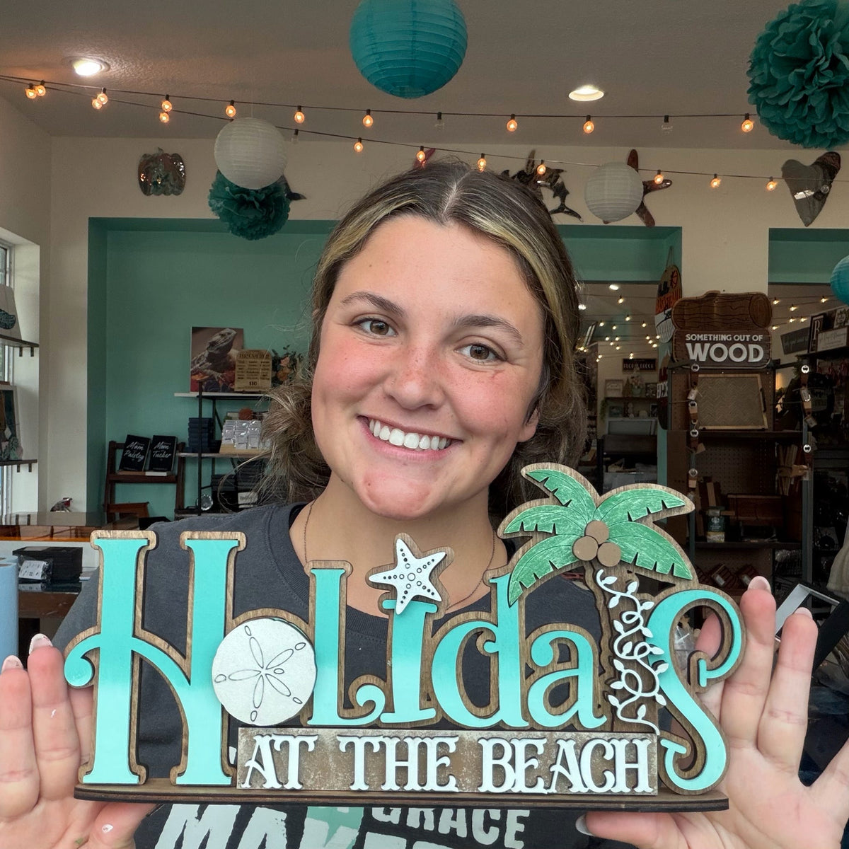 A smiling woman holds a decorative wooden sign that reads 'Holidays at the Beach' with turquoise lettering, a palm tree, and starfish motifs in a craft store setting.