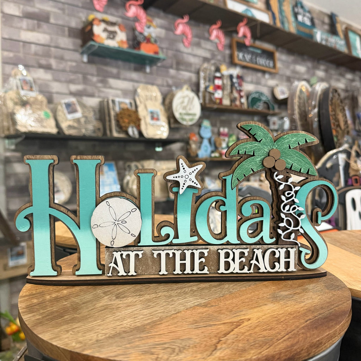 A decorative wooden sign reading 'Holidays at the Beach' in turquoise and white lettering, with a palm tree, starfish, and sand dollar motif, displayed on a round wooden table in a shop interior.