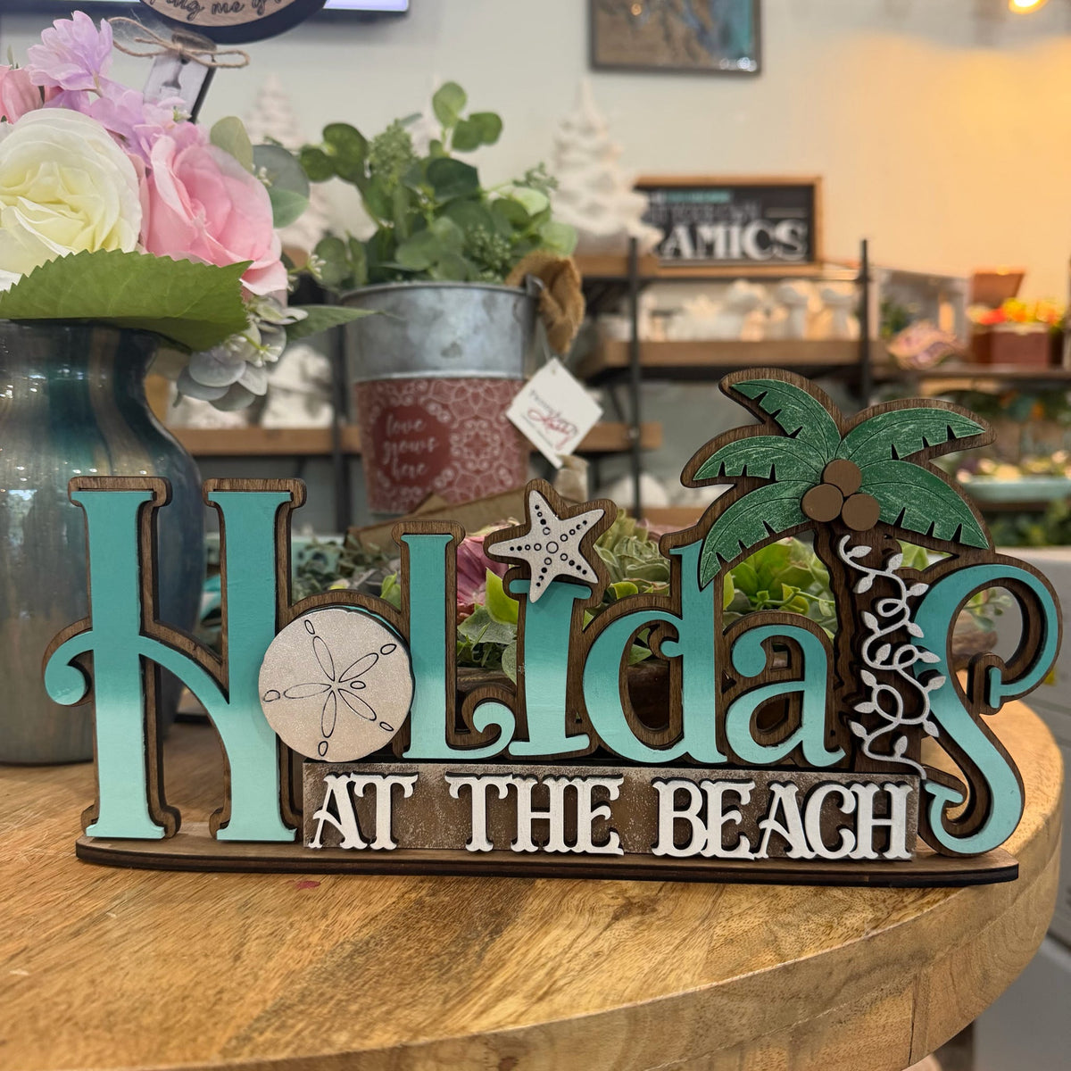 A decorative wooden sign reading 'Holidays at the Beach' in turquoise and white lettering, with a green palm tree and starfish accents, displayed on a wooden table in a shop setting.