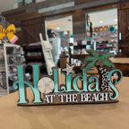 A decorative wooden sign reading 'Holidays at the Beach' with turquoise lettering, a palm tree, starfish, and sand dollar accents, displayed on a tabletop in a store interior.