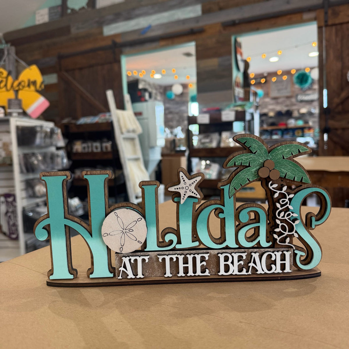 A decorative wooden sign reading 'Holidays at the Beach' with turquoise lettering, a palm tree, starfish, and sand dollar accents, displayed on a tabletop in a store interior.