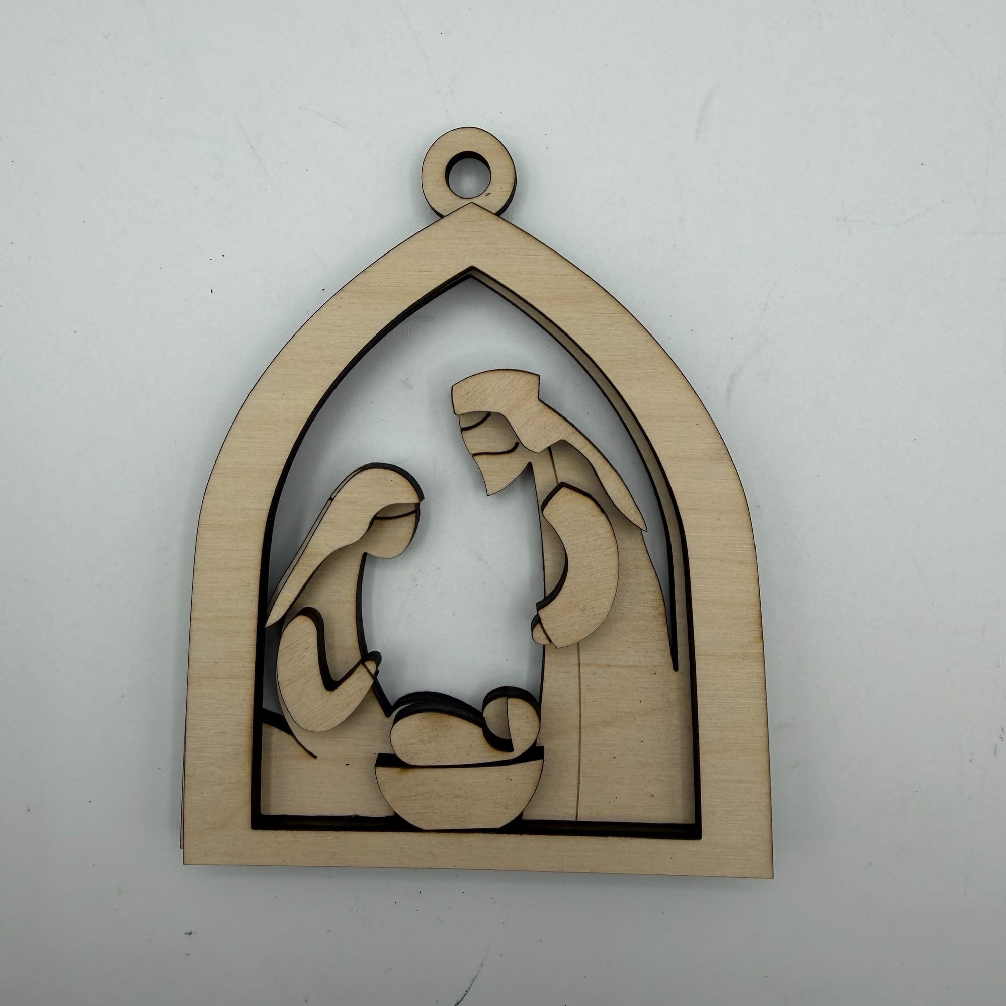 A laser-cut wooden nativity scene ornament featuring Mary, Joseph, and baby Jesus in a stable arch shape, shown on a plain light gray background.
