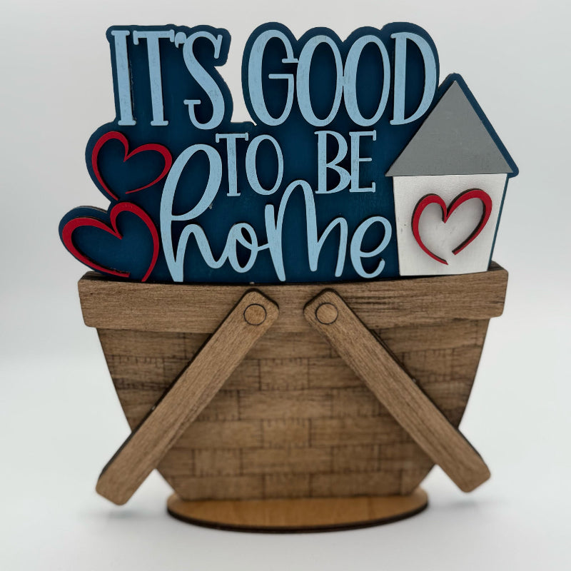 Decorative sign with 'It's Good to Be Home' text, hearts, and a house on a wooden base.