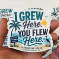 A hand holding a die-cut sticker that reads 'I GREW Here, YOU FLEW Here.' with tropical beach artwork including palm trees, a sun, waves, and a bird.