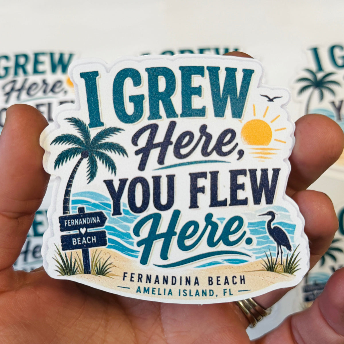 A hand holding a die-cut sticker that reads 'I GREW Here, YOU FLEW Here.' with tropical beach artwork including palm trees, a sun, waves, and a bird.