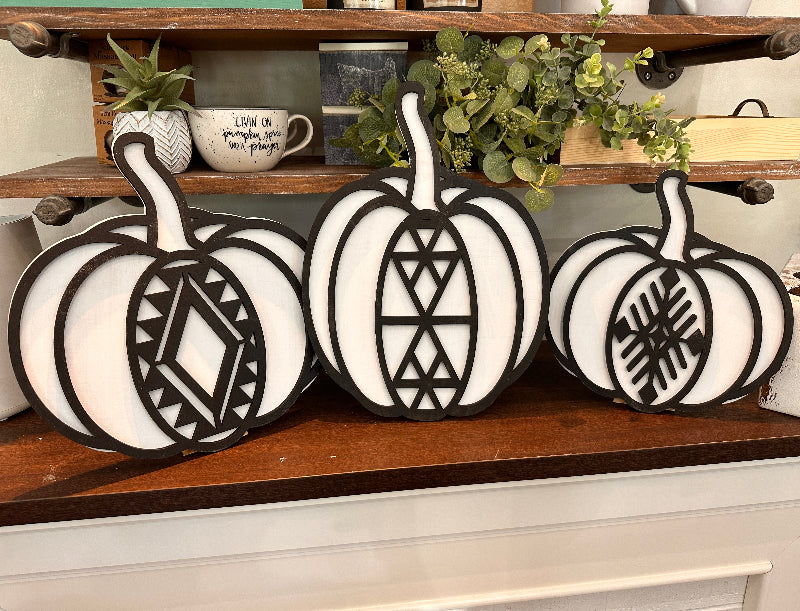 Three white pumpkins with geometric boho patterns laser cut on them, displayed on a wooden surface.