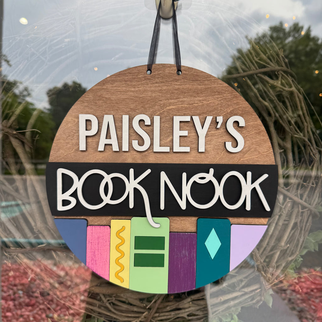 Decorative sign with 'Paisley's Book Nook' text and colorful design, hanging outdoors.