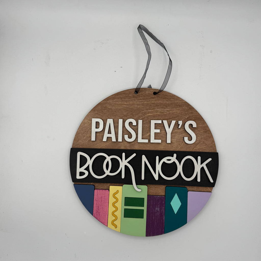 Round wooden sign with 'Paisley's Book Nook' text and colorful design on a white background