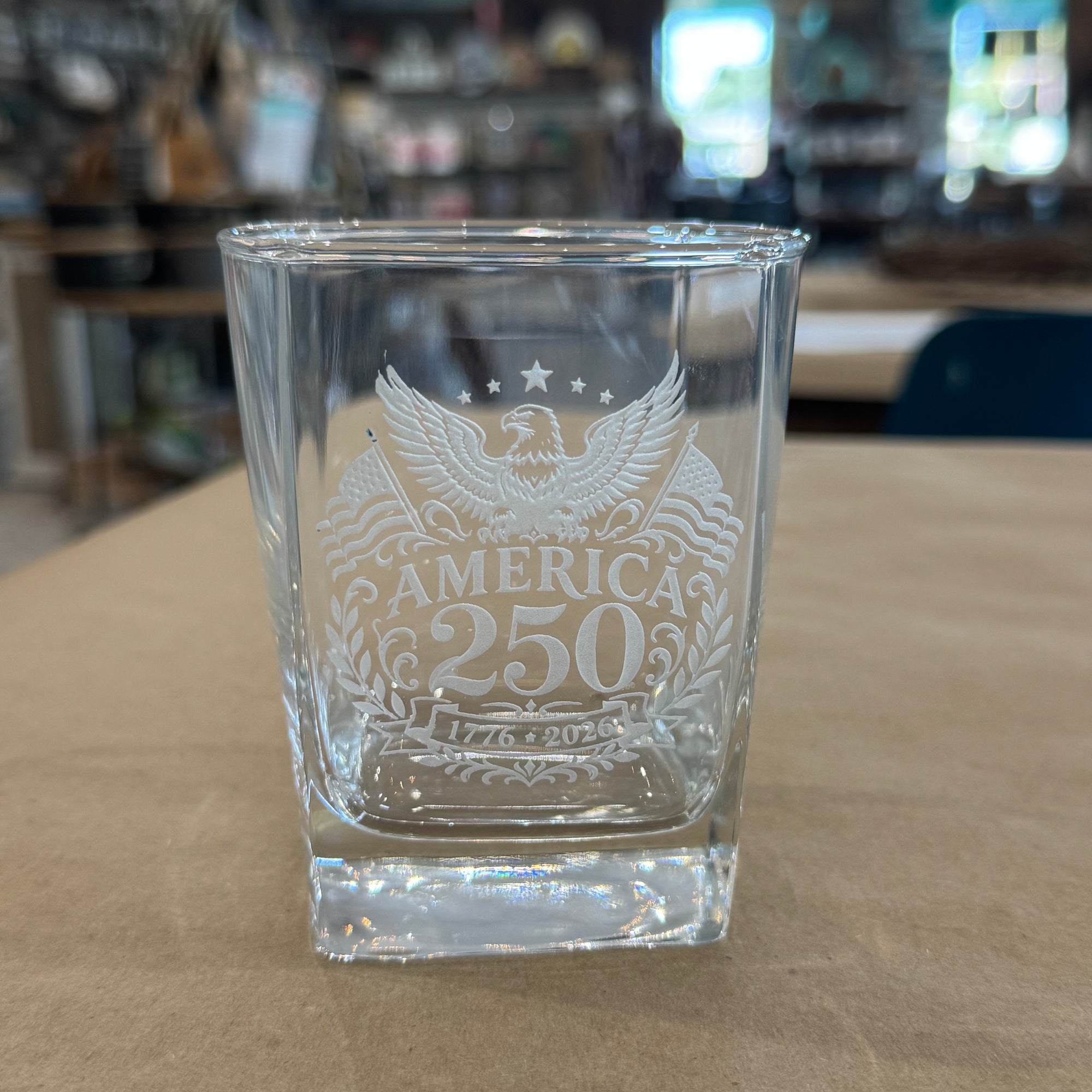 Clear glass shot glass with engraved design of an eagle and 'America 250' text on a wooden surface.