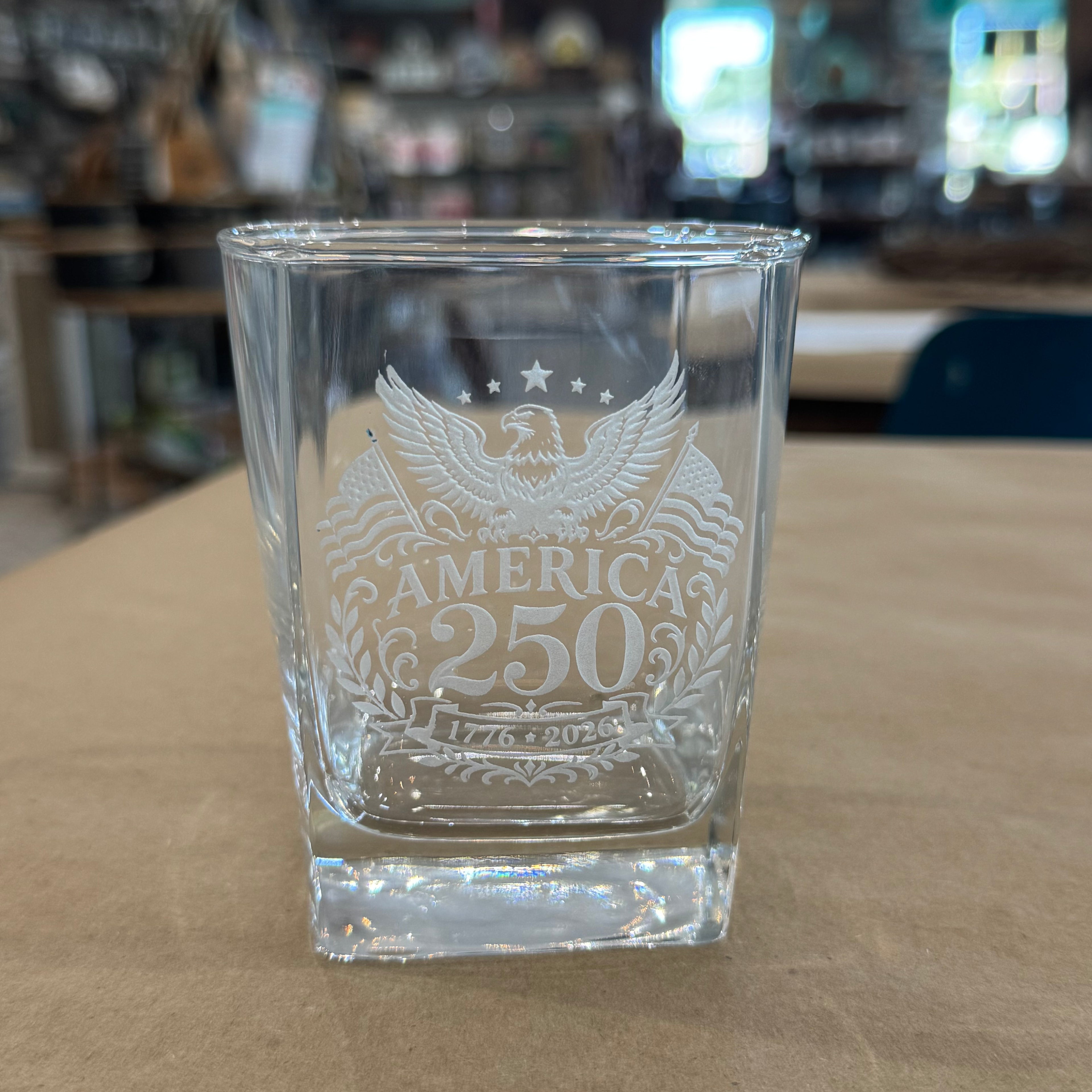 Clear glass shot glass with engraved design of an eagle and 'America 250' text on a wooden surface.