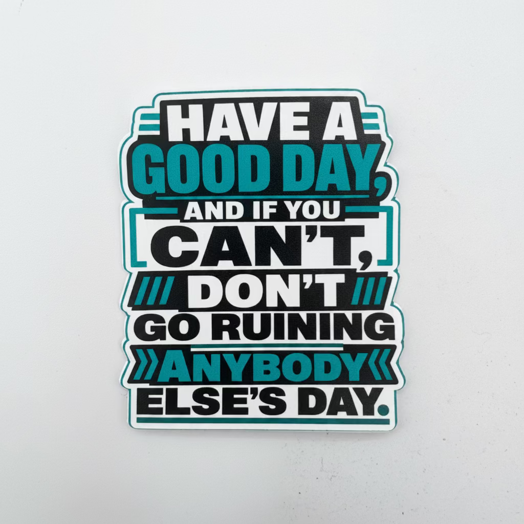 A die-cut vinyl sticker with a motivational quote, displayed on a light gray background. The sticker features a black background with white and teal text reading 'HAVE A GOOD DAY, AND IF YOU CAN'T, DON'T GO RUINING ANYBODY ELSE'S DAY.