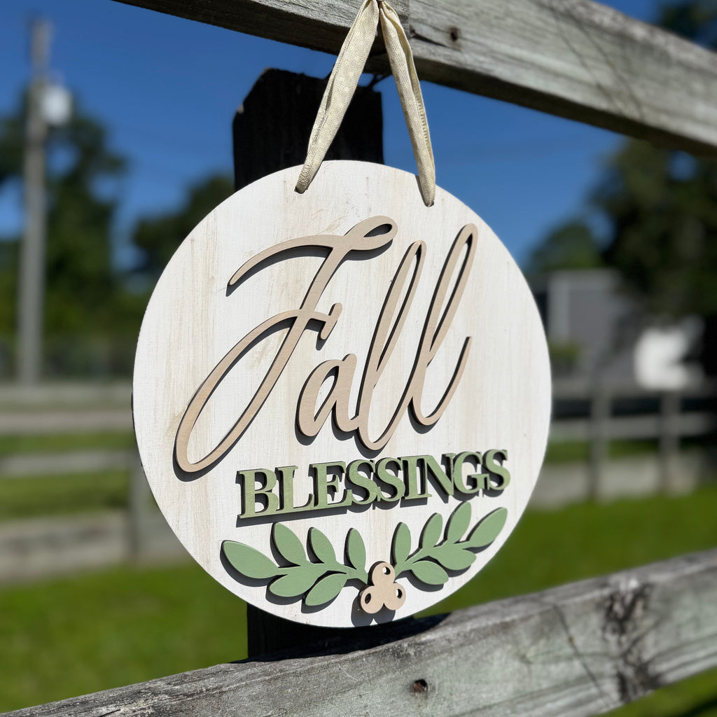 Round wooden hanging sign with a distressed white finish, featuring the words 'Fall Blessings' in beige and green lettering, with a green leaf design at the bottom, hanging from a wooden fence outdoors.