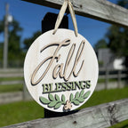Round wooden hanging sign with a distressed white finish, featuring the words 'Fall Blessings' in beige and green lettering, with a green leaf design at the bottom, hanging from a wooden fence outdoors.