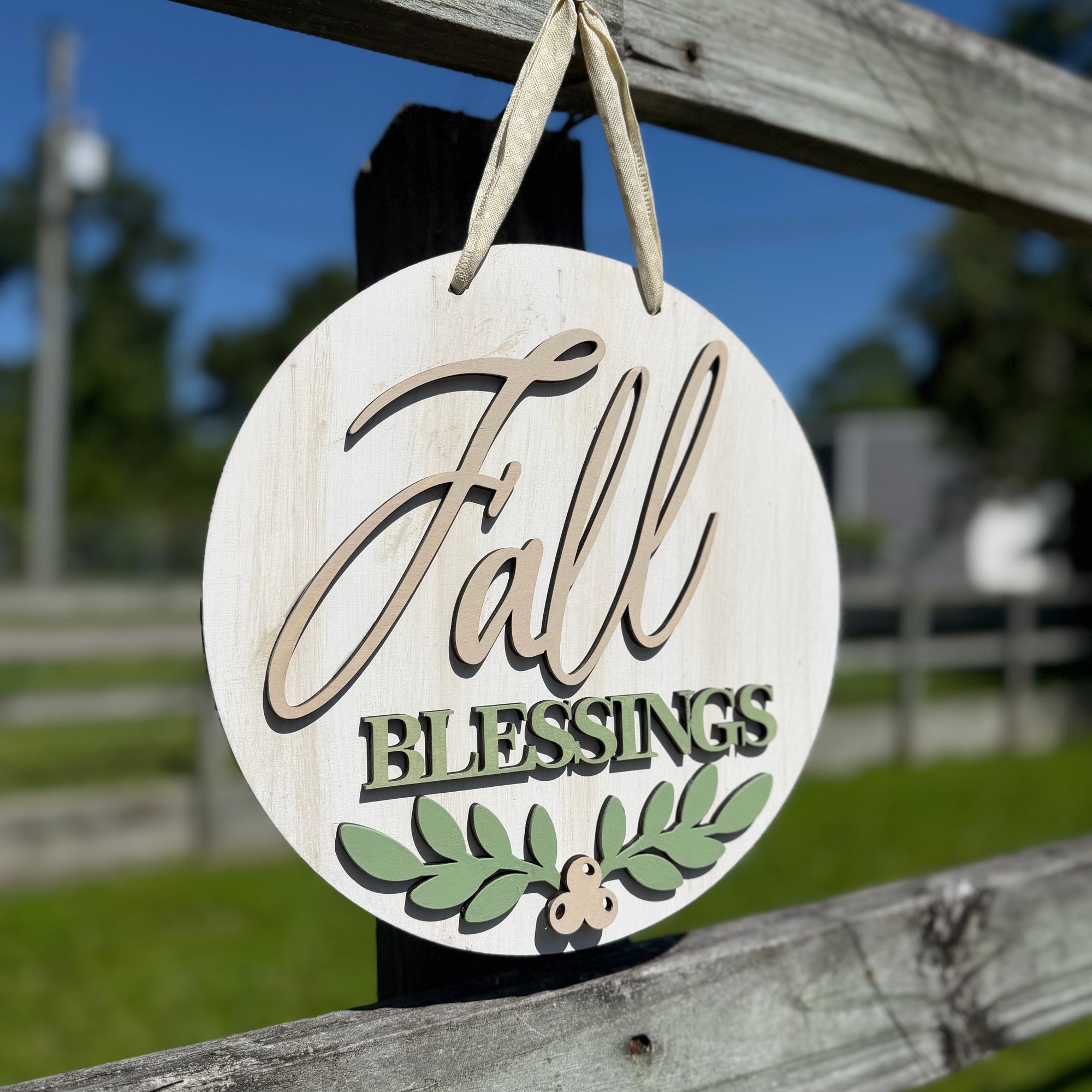 Round wooden hanging sign with a distressed white finish, featuring the words 'Fall Blessings' in beige and green lettering, with a green leaf design at the bottom, hanging from a wooden fence outdoors.