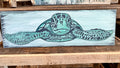 A wall-mounted wooden plank art piece with a turtle design painted in a beachy, watercolor style.