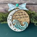 Decorative wooden ornament with wave design and text, tied with a ribbon on a green surface.